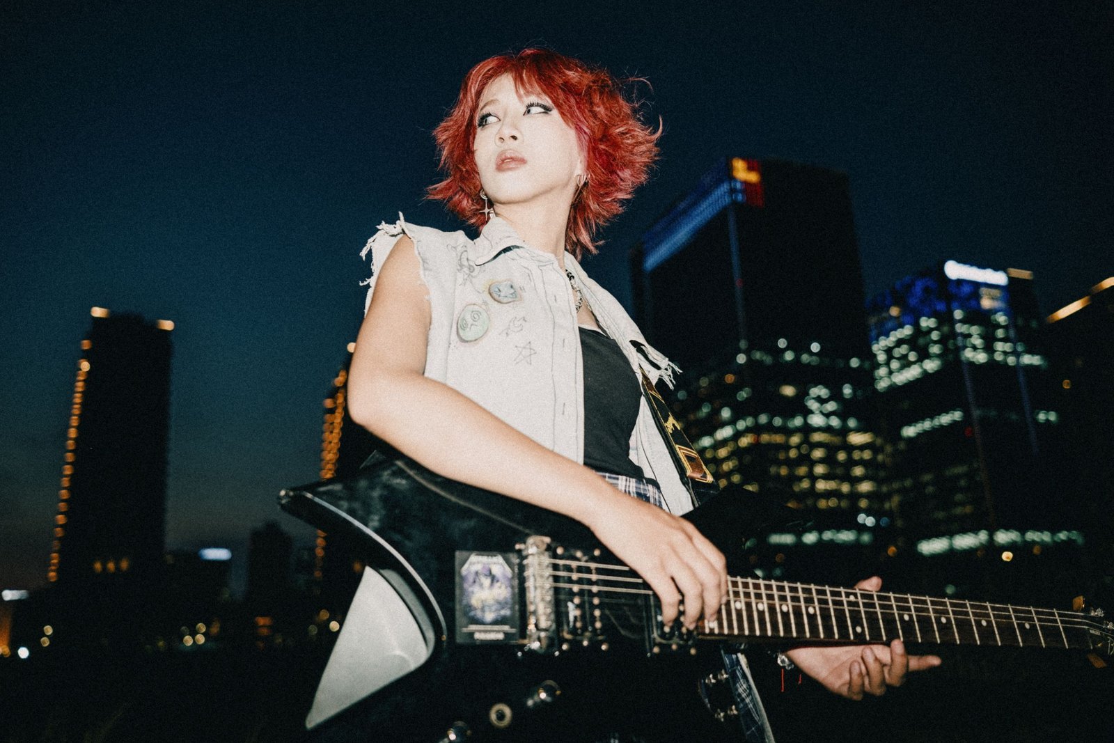 Donne e Chitarra: la Storia Nascosta dello Strumento più Amato del Rock 1 A woman with dyed hair plays an electric guitar against a city night skyline, showing urban rock vibes.
